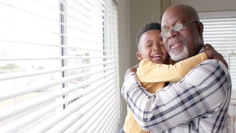 Glücklicher-Afroamerikanischer-Enkel-Und-Großvater,-Der-Sich-Zu-Hause-Am-Fenster-Umarmt,-Zeitlupe