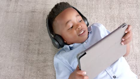 Happy-african-american-boy-in-headphones-lying-on-floor,-using-tablet-at-home,-slow-motion