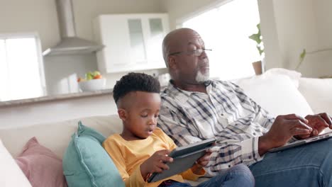 Abuelo-Afroamericano-Con-Nieto-Usando-Tableta-Y-Computadora-Portátil-En-El-Sofá-En-Casa,-Cámara-Lenta