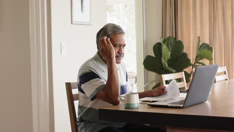 Biracial-senior-man-doing-paperwork-and-using-laptop-at-home,-slow-motion