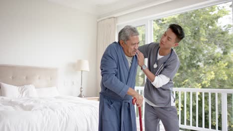 Diverse-male-doctor-helping-senior-male-patient-using-walking-stick-at-home,-slow-motion