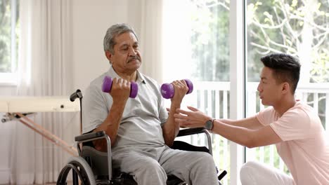 Diverse-male-physiotherapist-advising-and-senior-man-in-wheelchair-using-dumbbells,-in-slow-motion