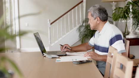 Biracial-senior-man-doing-paperwork-and-using-laptop-at-home,-slow-motion