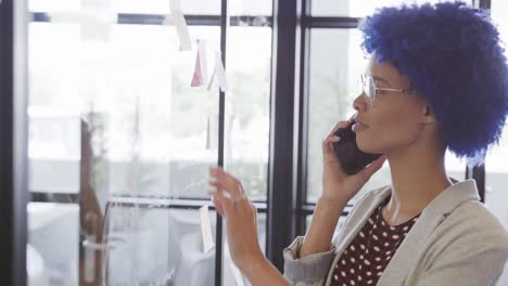 Portrait-of-biracial-businesswoman-making-notes-on-glass-wall-and-talking-on-smartphone,-slow-motion