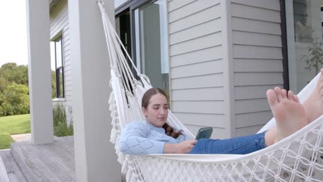 Happy-caucasian-woman-lying-in-hammock-and-using-smartphone-in-garden-in-slow-motion