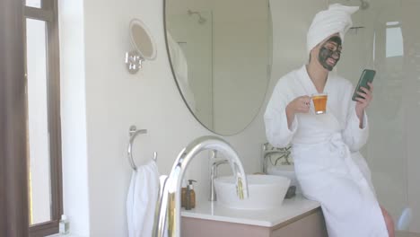 Fröhliche-Kaukasische-Frau-Mit-Maske,-Die-Tee-Trinkt-Und-Ihr-Smartphone-Im-Badezimmer-In-Zeitlupe-Benutzt