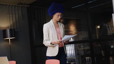 Biracial-casual-businesswoman-with-blue-afro-holding-documents-and-using-laptop,-slow-motion