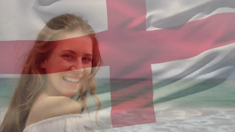 Animation-of-flag-of-england-waving-over-beautiful-smiling-biracial-woman-standing-at-beach