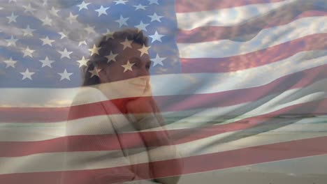 Animation-of-flag-of-america-waving-over-senior-caucasian-woman-feeling-cold-while-walking-on-beach