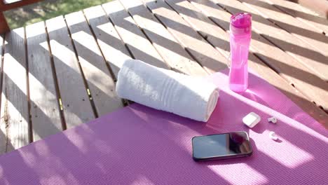 Handtuch,-Flasche-Wasser,-Smartphone-Und-Kopfhörer-Liegen-Auf-Yogamatte-Auf-Sonniger-Terrasse,-Zeitlupe