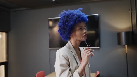 Portrait-of-happy-biracial-businesswoman-with-blue-afro-holding-glasses-in-meeting-room,-slow-motion