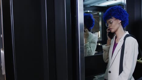 Biracial-businesswoman-with-blue-afro-talking-on-smartphone-and-using-elevator,-slow-motion