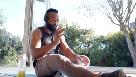 African-american-man-resting-on-mat-using-smartphone-and-headphones-at-sunny-home,-slow-motion