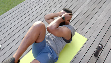 Focused-african-american-man-exercising-on-mat-with-bottle-of-water-in-garden,-slow-motion
