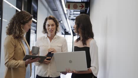 Diverse-female-colleagues-in-discussion-using-laptop-and-tablet-in-office-corridor,-slow-motion