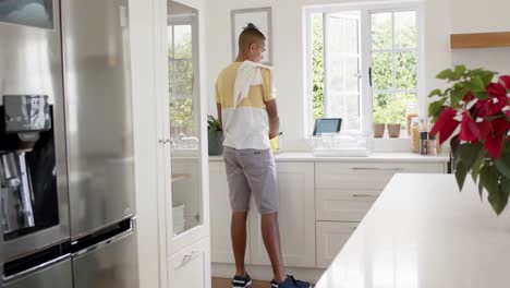 Biracial-man-washing-dishes-and-using-tablet-in-bright-kitchen,-slow-motion