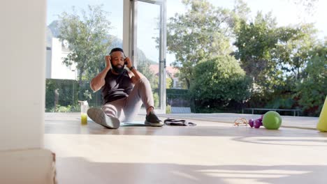 African-american-man-resting-on-mat-using-smartphone-and-headphones-at-sunny-home,-slow-motion
