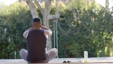 African-american-man-practicing-yoga-on-mat-with-bottle-of-juice-in-garden,-slow-motion