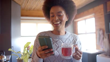 Feliz-Mujer-Afroamericana-Usando-Un-Teléfono-Inteligente-Y-Bebiendo-Té-En-Una-Cocina-Soleada,-En-Cámara-Lenta