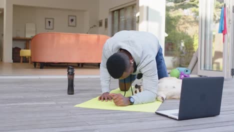 African-american-man-with-dog-exercising-on-mat-with-laptop-at-home,-slow-motion