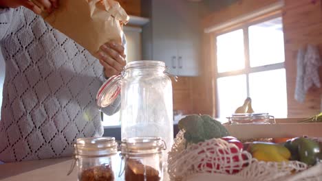 Mid-section-of-african-american-woman-pouring-pasta-into-jar-in-sunny-kitchen,-in-slow-motion