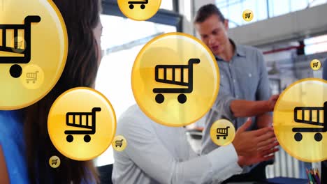 Animation-of-shopping-cart-icons-over-diverse-female-looking-at-coworkers-discussing-reports