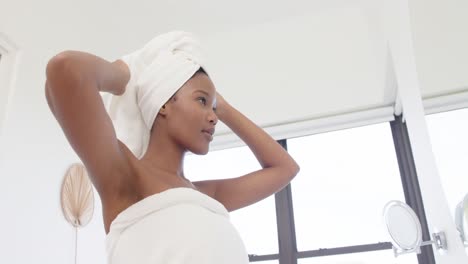 Happy-biracial-woman-with-towel-on-head-looking-in-mirror-in-bathroom,-slow-motion