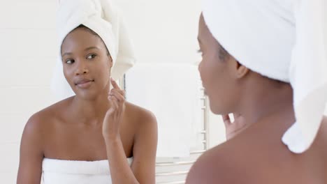 Happy-biracial-woman-with-towel-on-head-looking-in-mirror-applying-cream-on-her-face,-slow-motion