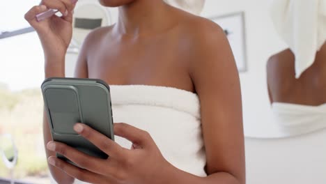 Happy-biracial-woman-with-towel-on-head-using-smartphone-and-brushing-teeth,-slow-motion