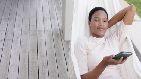 Feliz-Mujer-Afroamericana-Tumbada-En-Una-Hamaca-En-La-Terraza,-Usando-Un-Teléfono-Inteligente,-Cámara-Lenta