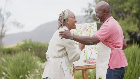 Happy-senior-african-american-couple-hugging-and-painting-on-wooden-easel-in-garden,-slow-motion