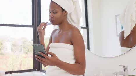 Happy-biracial-woman-with-towel-on-head-using-smartphone-and-brushing-teeth,-slow-motion