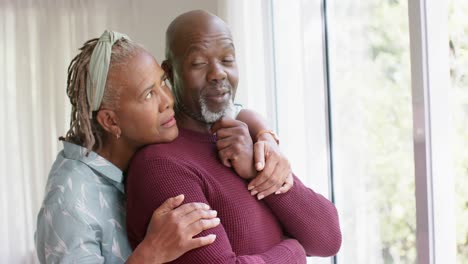 Nachdenkliches-Afroamerikanisches-Seniorenpaar,-Das-Sich-Zu-Hause-Umarmt-Und-Aus-Dem-Fenster-Schaut,-Zeitlupe