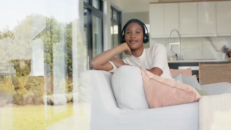 Feliz-Mujer-Afroamericana-Sentada-En-Un-Sofá-Usando-Auriculares,-Mirando-Por-La-Ventana,-Cámara-Lenta