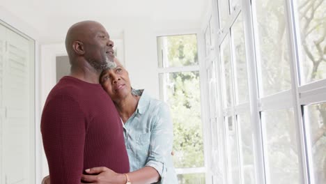 Nachdenkliches-älteres-Afroamerikanisches-Paar,-Das-Sich-Zu-Hause-Umarmt-Und-Aus-Dem-Fenster-Schaut,-Zeitlupe