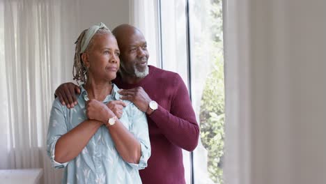 Reflexiva-Pareja-De-Ancianos-Afroamericanos-Abrazándose-Y-Mirando-Por-La-Ventana-En-Casa,-Cámara-Lenta