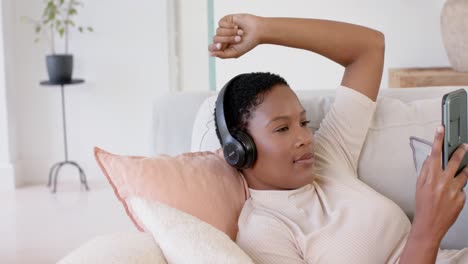 Glückliche-Afroamerikanische-Frau,-Die-Auf-Dem-Sofa-Liegt,-Kopfhörer-Trägt-Und-Smartphone-Benutzt,-Zeitlupe