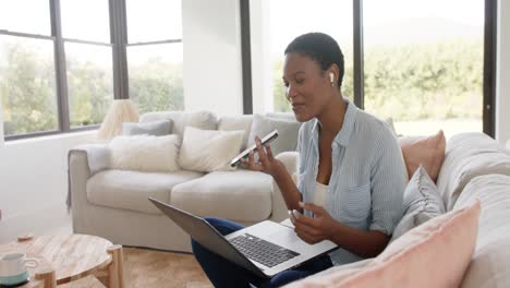 Fröhliche-Afroamerikanische-Frau-Sitzt-Auf-Dem-Sofa-Und-Spricht-Auf-Dem-Smartphone-Und-Benutzt-Einen-Laptop,-Zeitlupe