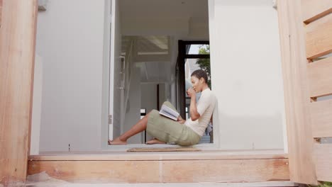 African-american-woman-sitting-in-doorway,-reading-book-and-drinking-tea,-slow-motion