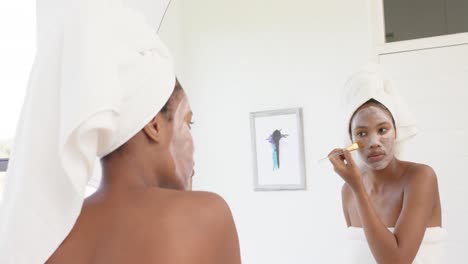 Focused-biracial-woman-with-towel-on-head-making-mask-on-face-in-bathroom,-slow-motion