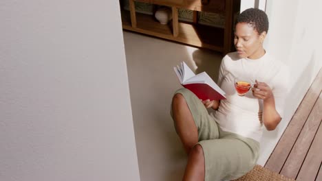 Afroamerikanische-Frau-Sitzt-In-Der-Tür,-Liest-Ein-Buch-Und-Trinkt-Tee,-Zeitlupe