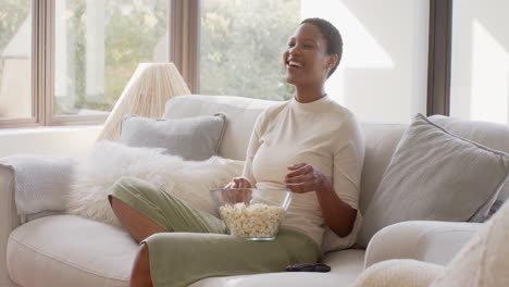 Feliz-Mujer-Afroamericana-Sentada-En-El-Sofá,-Viendo-Televisión-Y-Comiendo-Palomitas-De-Maíz,-Cámara-Lenta