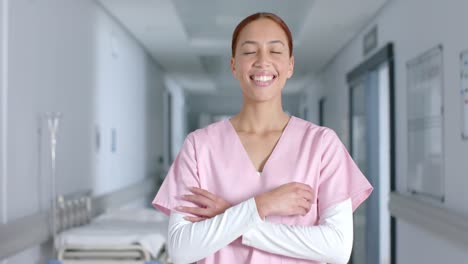 Portrait-of-happy-biracial-female-doctor-wearing-scrubs,-smiling-in-corridor,-slow-motion