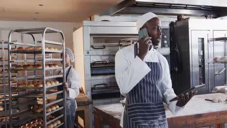 Diverse-bakers-in-bakery-kitchen,-talking-on-smartphone-and-holding-bakery-sheets-in-slow-motion