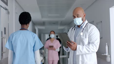 Portrait-of-biracial-male-doctor-wearing-face-mask,-using-tablet-in-corridor,-slow-motion