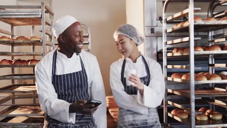 Fröhliche,-Vielfältige-Bäcker,-Die-In-Der-Bäckereiküche-Arbeiten-Und-Ihr-Smartphone-In-Zeitlupe-Nutzen