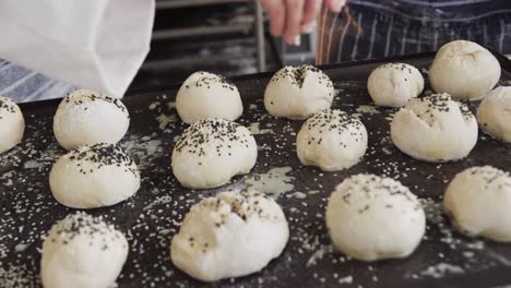 Diverse-bakers-working-in-bakery-kitchen,-sprinkling-poppy-seeds-on-rolls-in-slow-motion