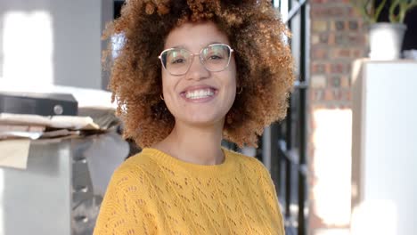 Portrait-of-biracial-young-businesswoman-with-afro-hair-wearing-glasses-and-smiling-in-office
