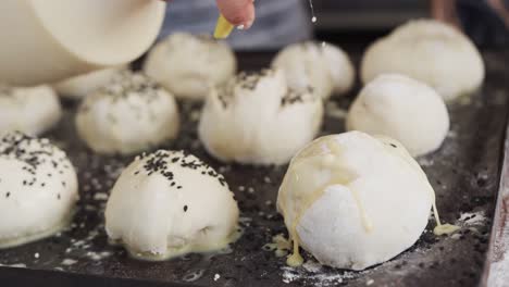 Asian-baker-working-in-bakery-kitchen,-spraying-and-sprinkling-poppy-seeds-on-rolls-in-slow-motion