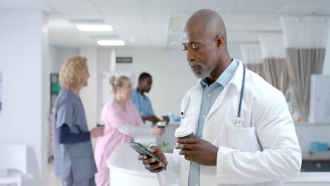 Glücklicher-Afroamerikanischer-Männlicher-Arzt,-Der-Im-Krankenhaus-Smartphone-Benutzt-Und-Kaffee-Trinkt,-Zeitlupe